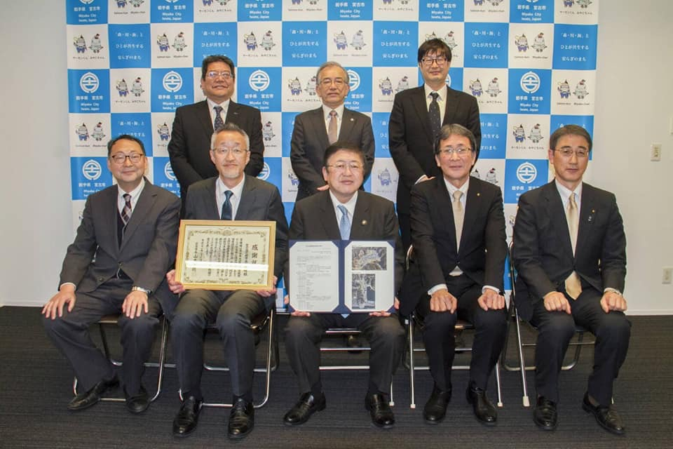 バックパネルの前で椅子に座り見開きのパネルのようなものを持つ市長を中心に賞状を持つ関係者の男性1名と3名の男性が並び、後列に3名の男性が立って並んでいる記念写真