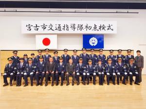「宮古市交通指導隊初点検式」と書かれた横断幕と日本国旗、宮古市の市町村旗が吊るされた会場で、前列に13名の関係者と制服を着た方々が椅子に座り、後列に15名の関係者と制服を着た方々が立って並んでいる記念写真
