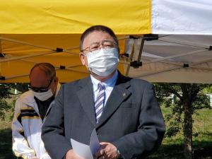 黄色と白のテントの下で作業をする様子の男性と、その前方で両手に資料を持ち顔を斜め前に向けている市長が写っている写真