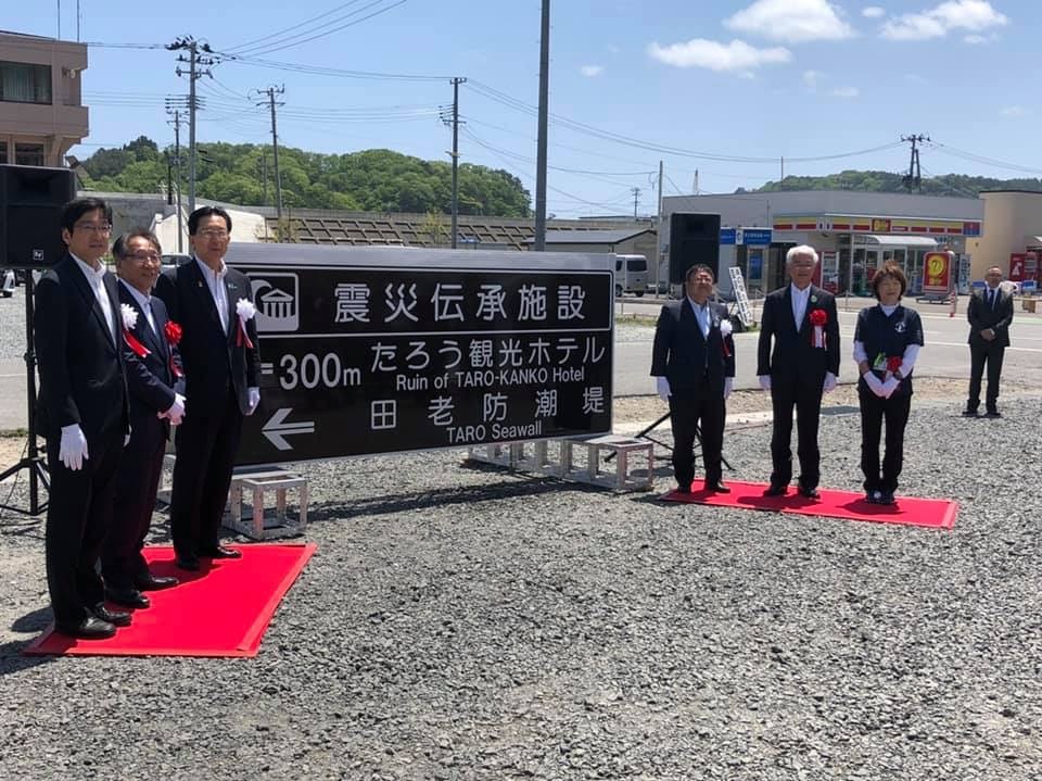 「震災伝承施設 たろう観光ホテル 田老防潮堤」と書かれた案内標識の左右に3名ずつ市長を含めた関係者が立っているお披露目式の様子の写真