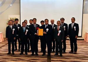 大きなスクリーンが設置された「令和4年度東北国道協議会総決起大会」会場で、谷藤裕明会長と東北地方整備局長の山本巧氏が要望書を一緒に持ち、市長を含め10名の関係者が横2列に並んでいる写真