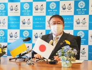 ウクライナと日本の柄のついた小さい国旗が斜めに交わるように置かれた茶色いテーブルの上に両手を置き、椅子に腰かけて斜め前を向いている様子の市長の写真