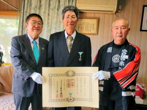 「瑞宝単光章」を受章された山本さんを中央に、賞状を持った市長と消防の関係者の男性が横一列に並んでいる写真