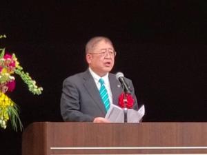 赤色の胸章リボンをつけた市長が演台のマイクを使いながら挨拶をしている写真