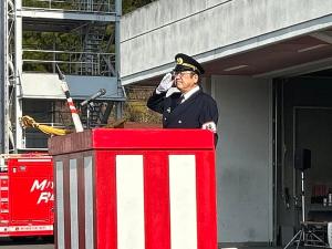 紅白で彩られた演台で敬礼をしている制服姿の市長を斜め横から写した消防出初式の写真