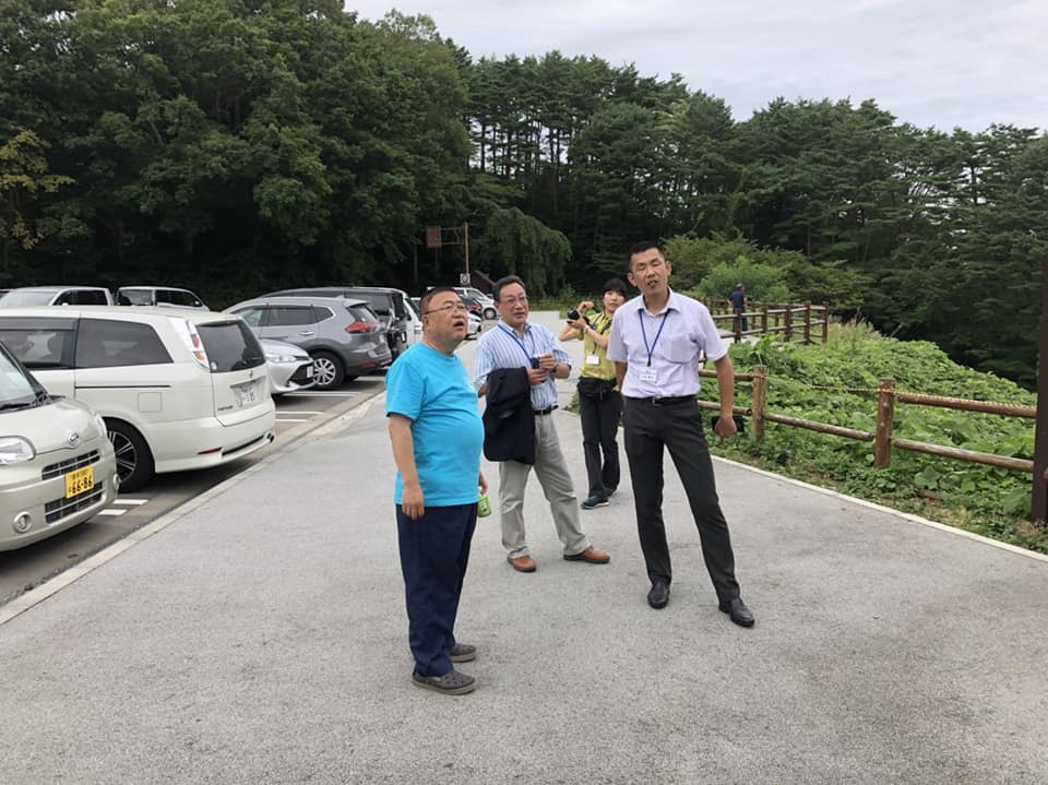 駐車場の脇で、立って話をする様子の青いTシャツを着た市長と2名の男性、カメラを構えた女性の写真