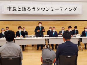 「市長と語ろうタウンミーティング」と書かれた横断幕が設置された会場で、市の関係者と参加者が向かい合って座り、中央の席から立ち上がった市長が挨拶を行っている写真