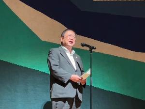ステージ上でスポットライトをあてられた市長が、スタンドマイクの前に立ち挨拶をしている様子の写真