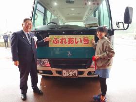新里地域バスのフロントにふれあい号と書かれた部分を 両端から白のリボン胸章をつけた市長とアイデアが採用され赤のリボン胸章をつけた 橋場悠さんが指さしている写真