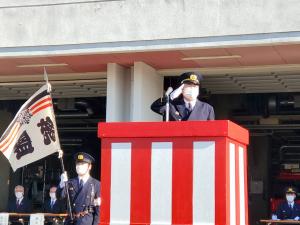 紅白幕の演説台に消防団の制服を着た男性が敬礼しており、その横に男性が消防旗を手に持っている写真