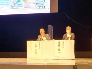 スクリーンが設置された舞台の上で白いテーブルクロスが引かれた机と椅子に座りマイクを持って話している様子の市長と、その隣に座る男性を写した写真