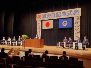 市民文化会館のステージに祝海の日記念式典と書かれた横断幕や日本国旗、宮古市の市旗等が掲げられ、出演者がステージ左右にそれぞれ設けられた席につき、中央にあるマイクが置かれた壇上で男性が話をしている写真