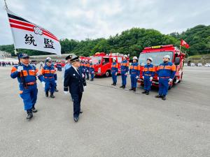 消防車の前に整列した消防団員の前を、紺色の制服に身を包んだ市長が「統監」と書かれた旗を持った団員の方々と一緒に歩いている消防団大演習の様子の写真