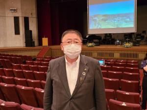景色の映像が映し出されたスクリーンをバックに、「室蘭市開港150年・市制施行100年記念式典」の会場の観客席に立つ市長の写真