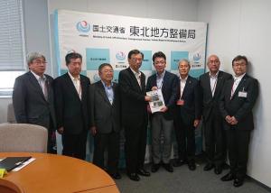 市長や国土交通省の関係者らスーツ姿の男性8名が横一列に並び中央に立つ2名が2人で要望書を持っている写真