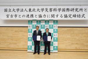 「国立大学法人東北大学災害科学国際研究と宮古市との連携と協力に関する協力締結式」と書かれた横断幕の下で賞状のようなものを前方に向け横に並ぶスーツを着た2人の男性の写真