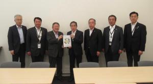 市長や公明党の関係者らスーツ姿の男性7名が横一列に並び中央に立つ2名が2人で要望書を持っている写真