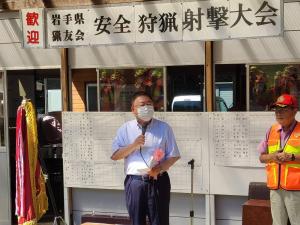 「歓迎 岩手県猟友会 安全狩猟射撃大会」と書かれ、優勝旗が置かれた会場で、右手にマイクを持ち挨拶を行っている市長の写真