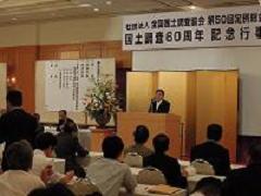国土調査60周年記念行事と書かれた吊り看板があり、金屏風を背景に演壇に立つ市長と、話を聞く参加者達を後ろから写した写真