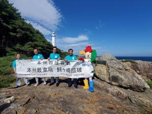 青空の下、魹ケ崎の岩場で「本州最東端魹ケ崎踏破」と書かれた横断幕をお揃い青のTシャツを着た市長と男性3名、マスコットキャラクターよんたんで持っている写真