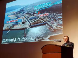 大きなスクリーンに「宮古港がより近い港に」と文字と写真が写され市長が説明をしている「いわてポートフォーラム2024」の写真