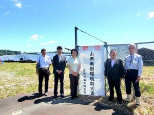 太陽光パネルが設置された敷地を囲むフェンスに「歓迎 山田美樹環境副大臣」と書かれた旗が掲げられ、山田環境副大臣と市長、3名の関係者が横一列に並んでいる写真