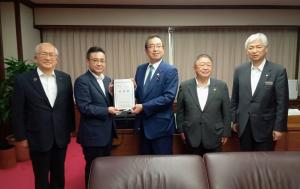 茶色い皮のソファーの後ろで、要望書を一緒に持つ男性2人、市長を含めた男性3人が横に並んでいる写真