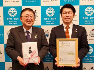 バックパネルの前で目録を持つ市長と、額縁を持つ男性が微笑んでいる写真