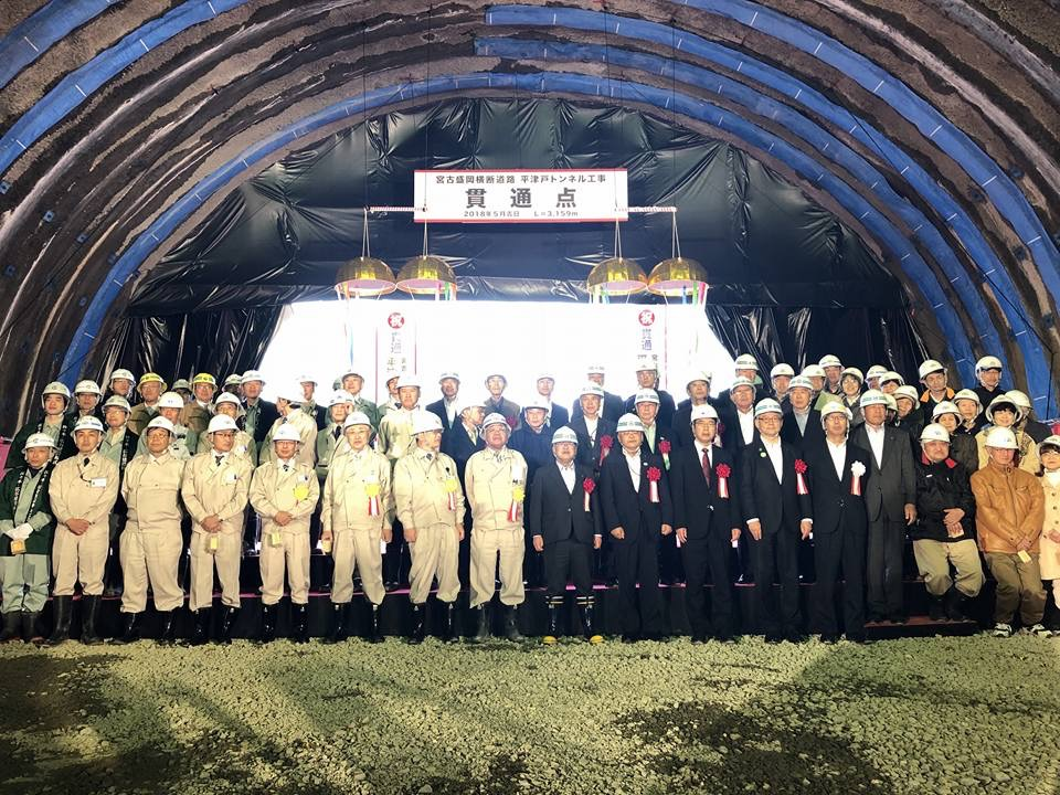 薄い黄色の作業着を着た方々や、スーツを着て胸章を着けた関係者の方々が3列になって並んで立つ記念写真