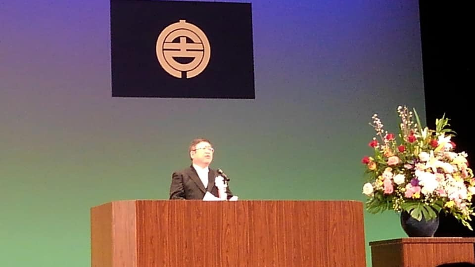 市旗が掲げられたステージの檀上に立つ市長が成人式会場で挨拶を行っている様子の写真