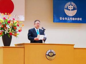 国旗、校旗が掲揚され、花が飾られ校章が貼られた演台で、市長が式辞用紙を見ながら祝辞を述べている写真