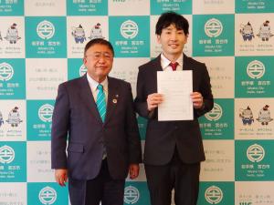 バックパネルの前で辞令を両手で持ち前方に掲げる男性と、その横に並ぶ市長を写した写真