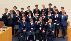 2名の高校生議員を中央に市長と関係者の男性が前列の椅子に座り、2列目と3列目に13名の高校生議員の皆さんと4名の関係者が立ちガッツポーズをしている記念写真