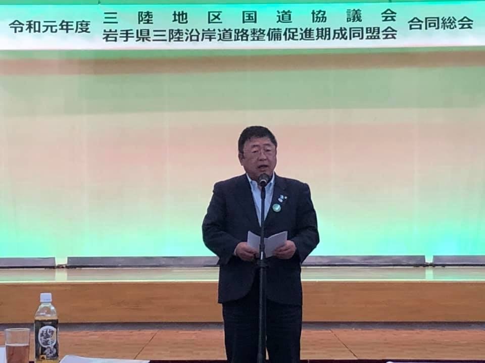「令和元年度 三陸地区国道協議会・岩手県三陸沿岸道路整備促進期成同盟会 合同総会」と書かれた横断幕が掲げられた会場で、中央に設置されたマイクスタンドの前に立ち挨拶を行っている写真