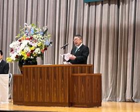 色鮮やかな花が活けられた大きな花瓶が置かれている演台の中心で手紙を見ながら話をしている様子の市長の写真
