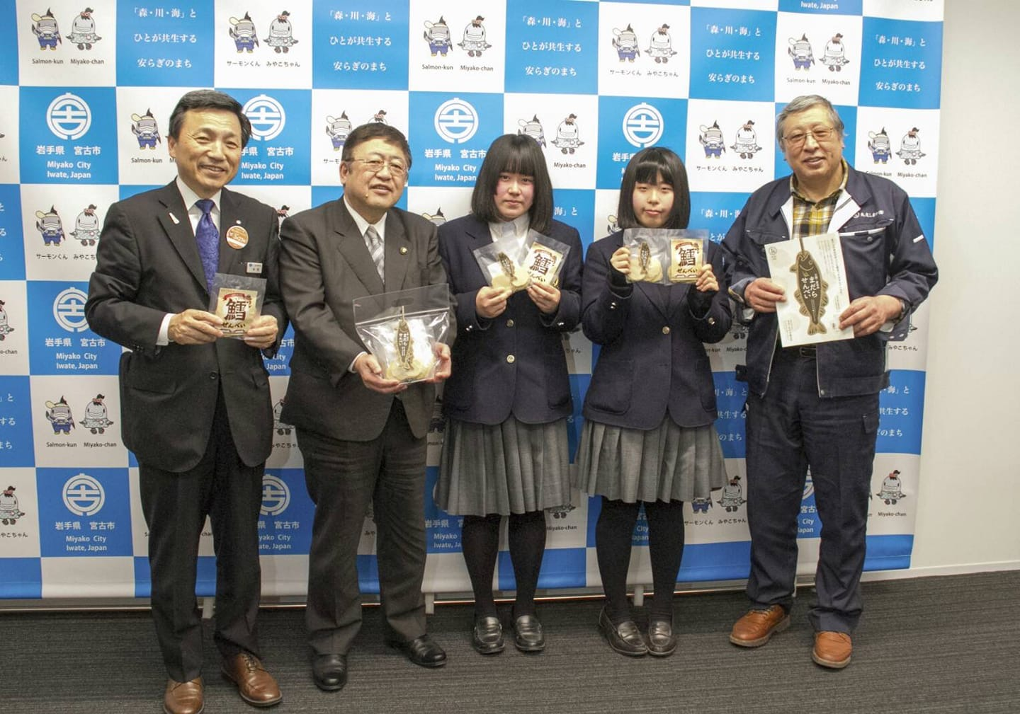 バックパネルの前で袋に入った三陸宮古のまだらせんべいの商品を持つ関係者の男性2名と市長、制服を着た女子高生の記念写真
