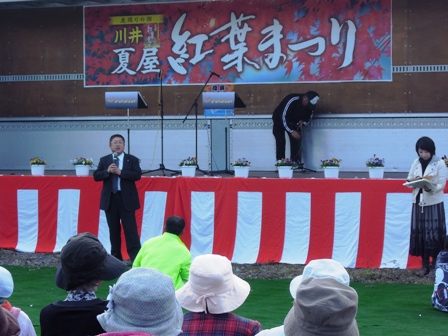 「夏屋紅葉まつり」の吊り看板が飾られた紅白幕の野外ステージ前中央で、市長がマイクを持ち観客の方に向けて話をしている様子の写真