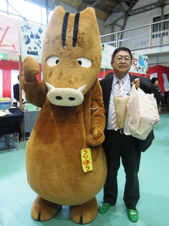 紙袋と袋いっぱいの品物を片手に持つ市長と、「うりぼう」の名札をつけたイノシシの着ぐるみが並んでいる写真