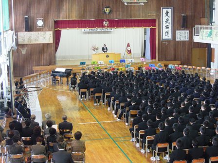 学校の体育館で椅子に座る生徒達と、式典に参加する来賓者が座っており、舞台の演壇で挨拶している写真