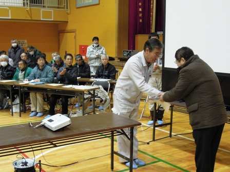 体育館に設置されたスクリーンの前で男性2名が握手しているのを長机に座った参加者たちが拍手している写真