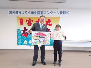 「宮古さけまつり小学生図画コンクール表彰式」と書かれた横断幕と大漁旗が飾られている前に立ち、賞状を手に持った小学生とカラフルに描かれた鮭の図画を手に持った市長の写真