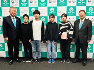 宮古市のバックパネルをバックに新里小学校の生徒4名が能登半島地震災害義援金の箱を持って市長と関係者の方にはさまれて記念撮影をしている写真