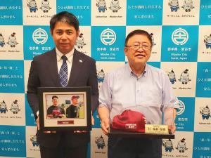 福井優也投手の写真が入った額縁を持つ男性と、ガラスケースに入った帽子を持つ市長がバックパネルの前で、立っている写真