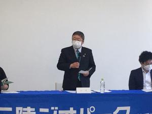 青い布が敷かれたテーブルの前に立ち、左手に資料、右手にマイクを持って話している様子の市長とその両側に座る関係者2人の写真