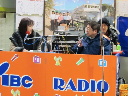 宮古市魚菜市場内で、ラジオの機材やコードが並べられた前のブースで、ラジオパーソナリティの女性とマイクを手に持ちながら会話する様子の写真