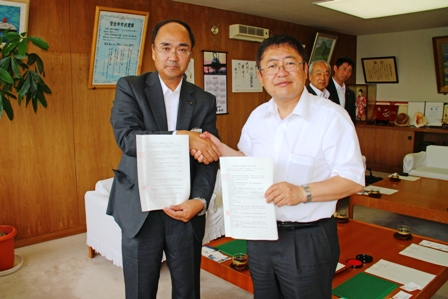 山本正徳市長と笠原治清事務局長が片手で握手をしもう片方の手で協定書を持ち記念撮影している写真