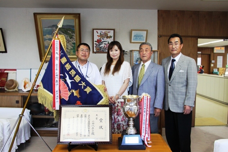 産経民謡大賞と書かれた優勝旗と賞状、優勝カップが飾られた後ろで男性3名と女性1名が並び記念撮影している写真