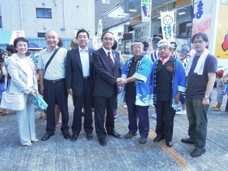 半被をきた市長とスーツをきた男性が両手で握手をし、その横に男性や女性が横に並び記念撮影している写真