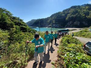 青空の下、後方に海が見える道を縦一列に並んで、お揃い青のTシャツを着た市長と男性3名がウォーキングポールを使って歩いている写真