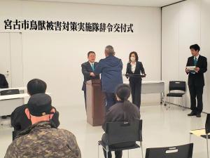 「宮古市鳥獣被害実施隊辞令交付式」と書かれた幕が貼られた会場で、他の参加者が座っている中、市長が演台で1名に辞令を交付している写真